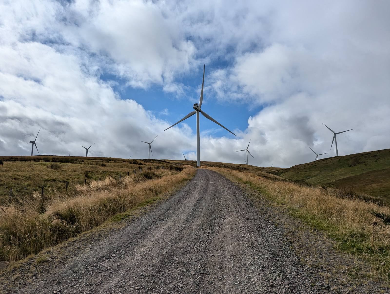 Onshore wind farm with multiple turbines on rolling Welsh hills, gravel access track leading to a centred turbine under bright cloudy sky