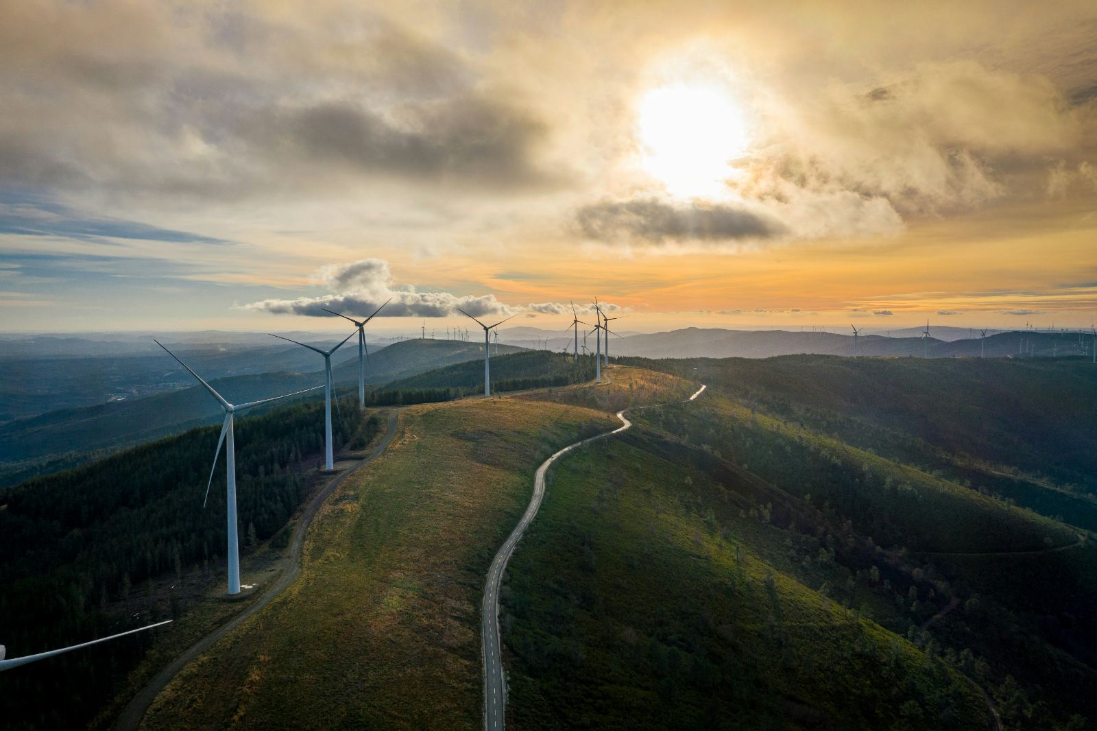 Aerial view of a ridge-top wind farm at sunset, single-track road threading along the spine of a forested mountain ridge with multiple turbines silhouetted against orange and grey cloud