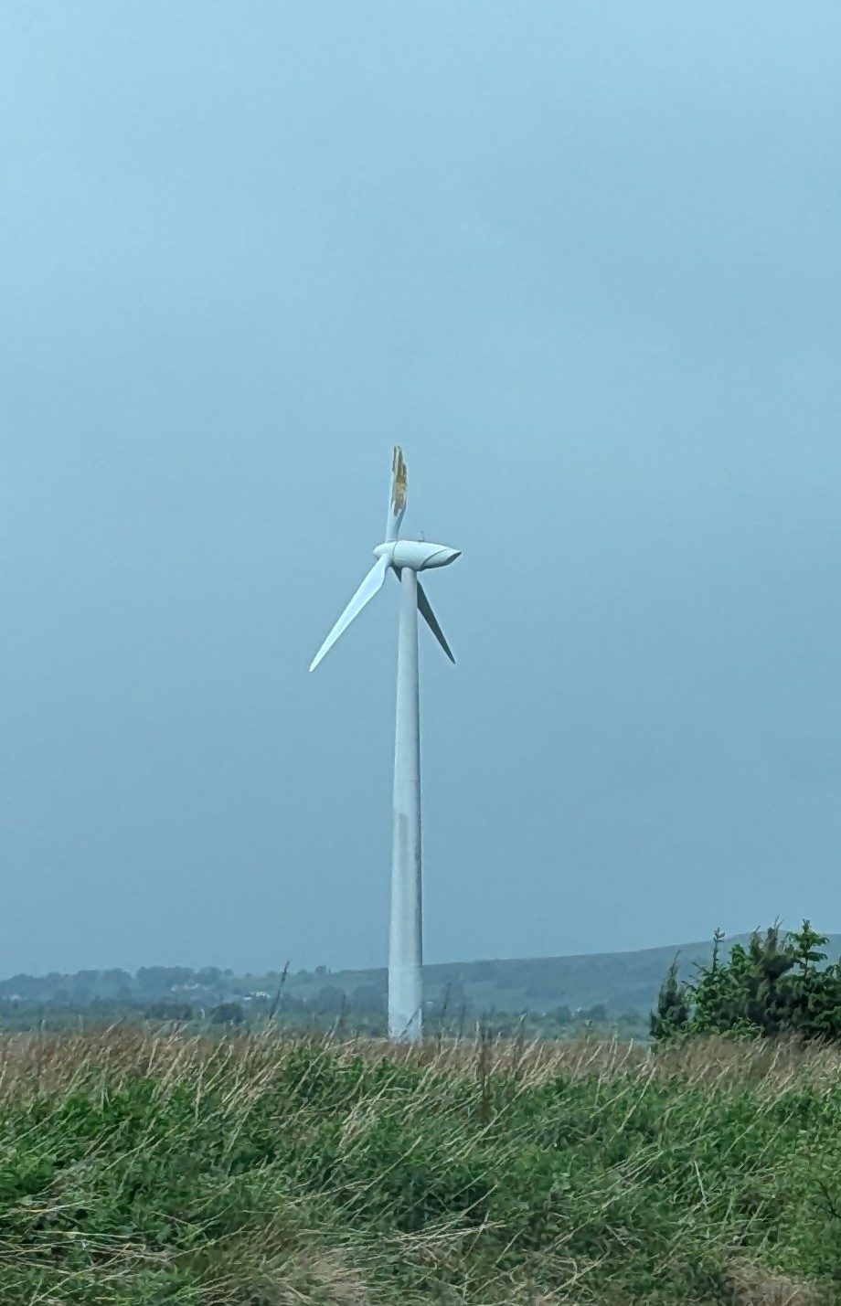 A wind turbine against an overcast sky with the tip of one blade visibly broken and splintered.