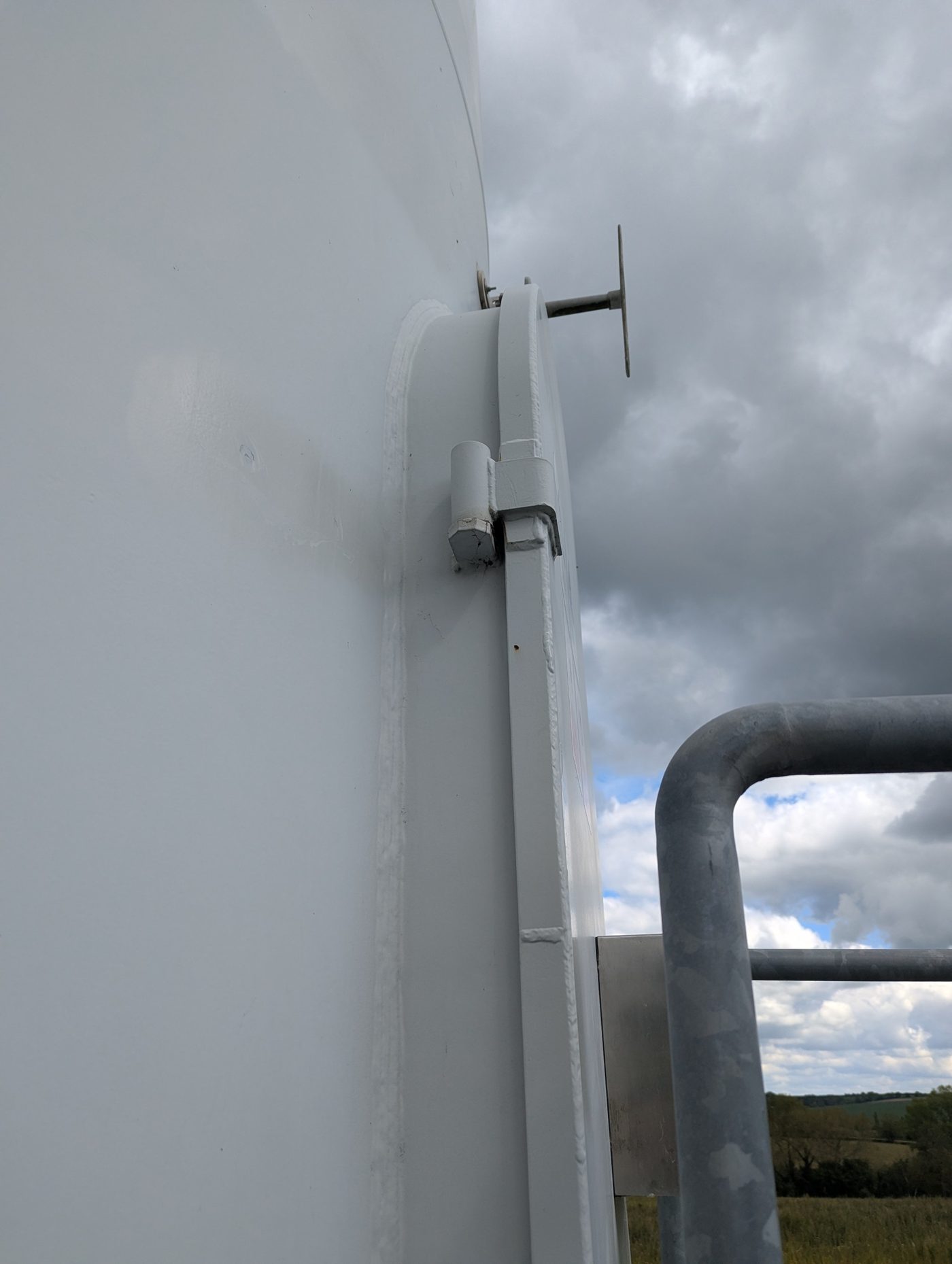 Close-up of the weld and hinge detail where a wind turbine tower door surround meets the tower shell, seen from below against a cloudy sky.